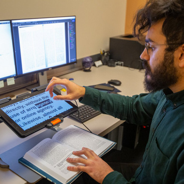 A person wearing glasses is sitting in a computer lab, reading a book with the help of an eletronic magnification aid.